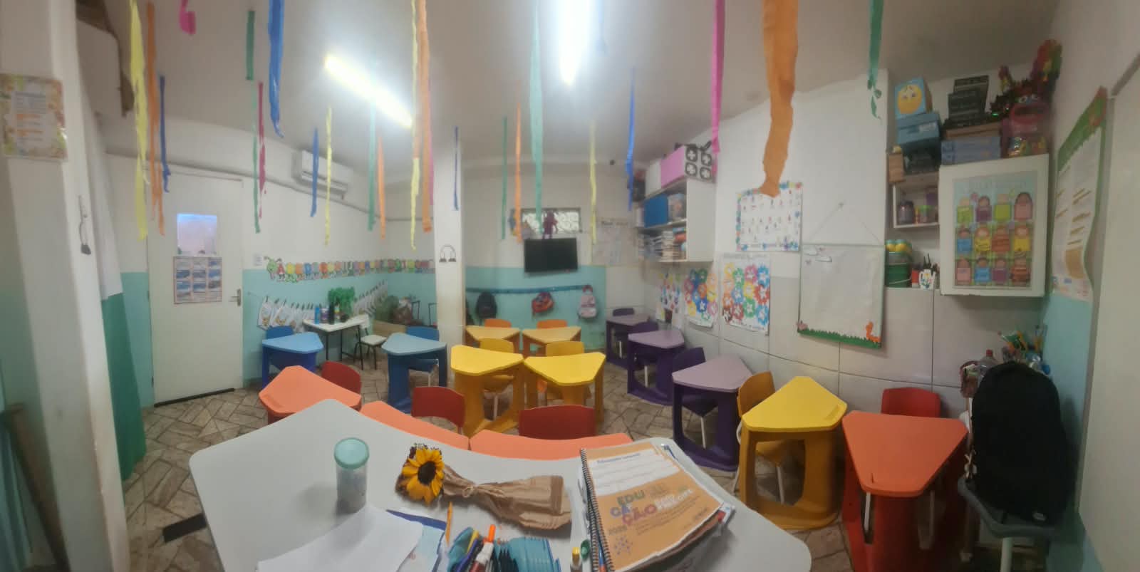 Sala da instituição com mesas e cadeiras infantis coloridas e materiais pedagógicos.