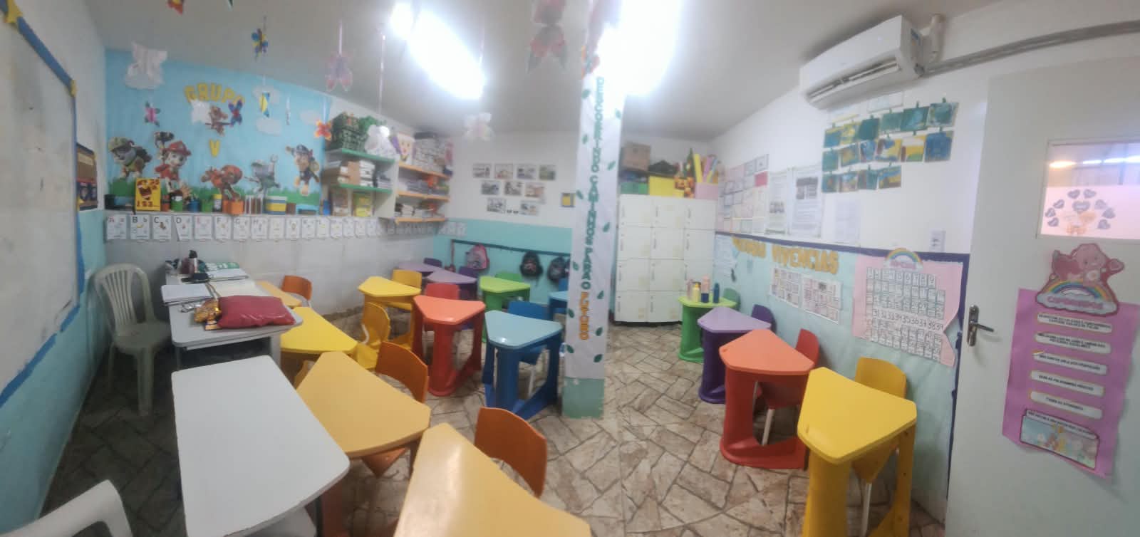 Sala de educação infantil com mesas coloridas, atividades nas paredes e materiais escolares.